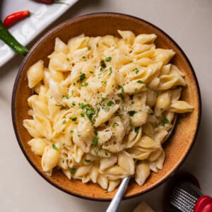 A bowl of creamy shell pasta topped with grated cheese and herbs sits on a table with a spoon. Nearby are green and red chili peppers on a white plate, a small cheese grater, and a cube of cheese.