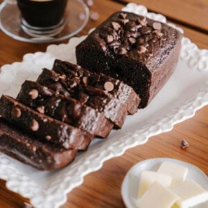 A sliced Chocolate Loaf Cake topped with chocolate chips is displayed on a white platter. Nearby, a cup of black coffee sits beside a small plate with butter slices and a red-handled butter knife.