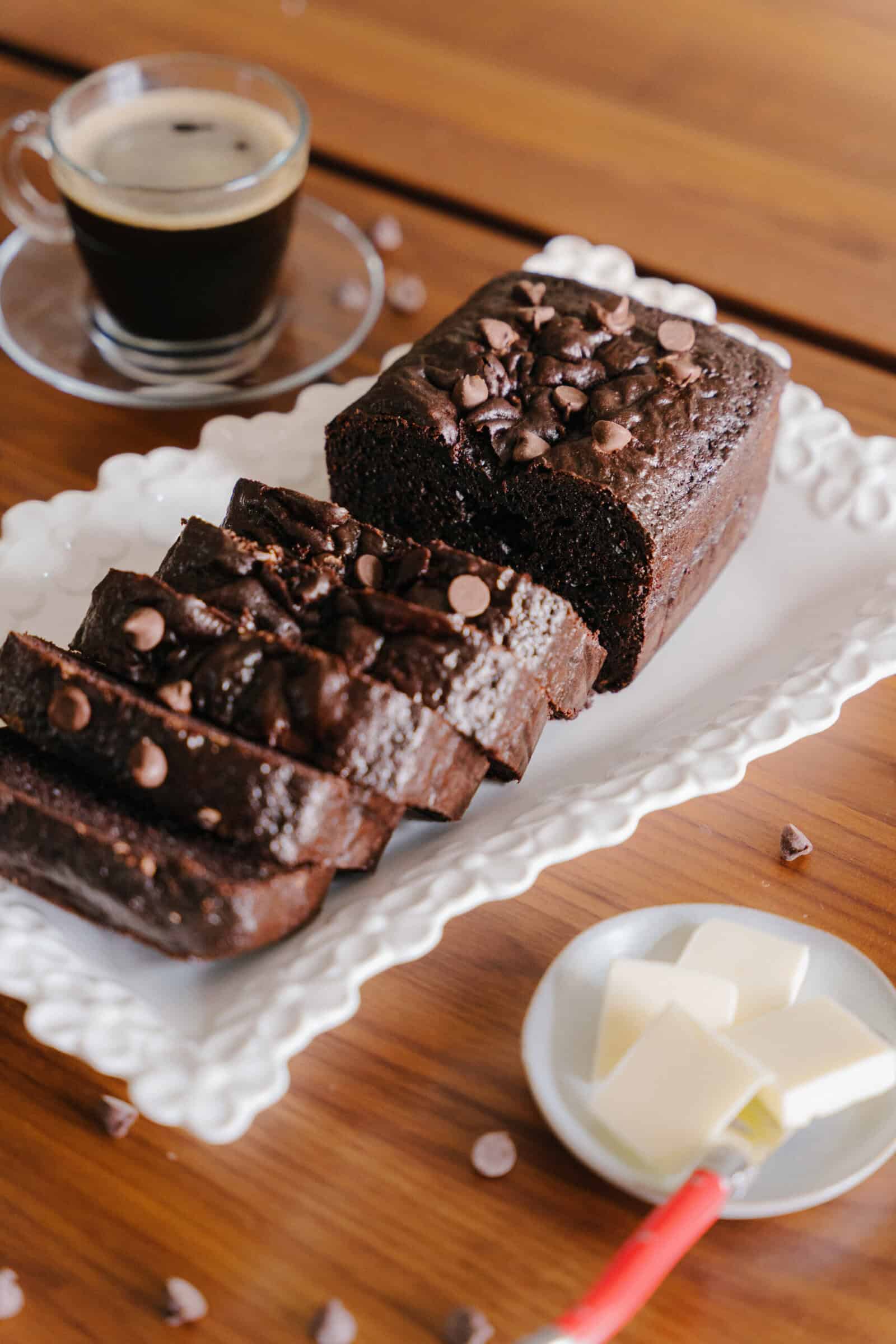 A sliced Chocolate Loaf Cake topped with chocolate chips is displayed on a white platter. Nearby, a cup of black coffee sits beside a small plate with butter slices and a red-handled butter knife.