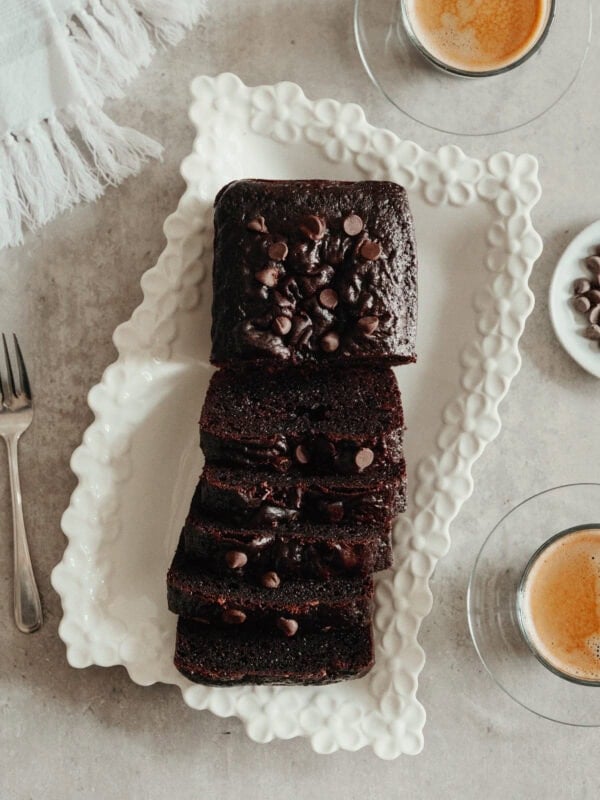 A sliced Chocolate Loaf Cake topped with chocolate chips is arranged on a decorative white platter, accompanied by two cups of espresso and a fork on a light-colored surface.