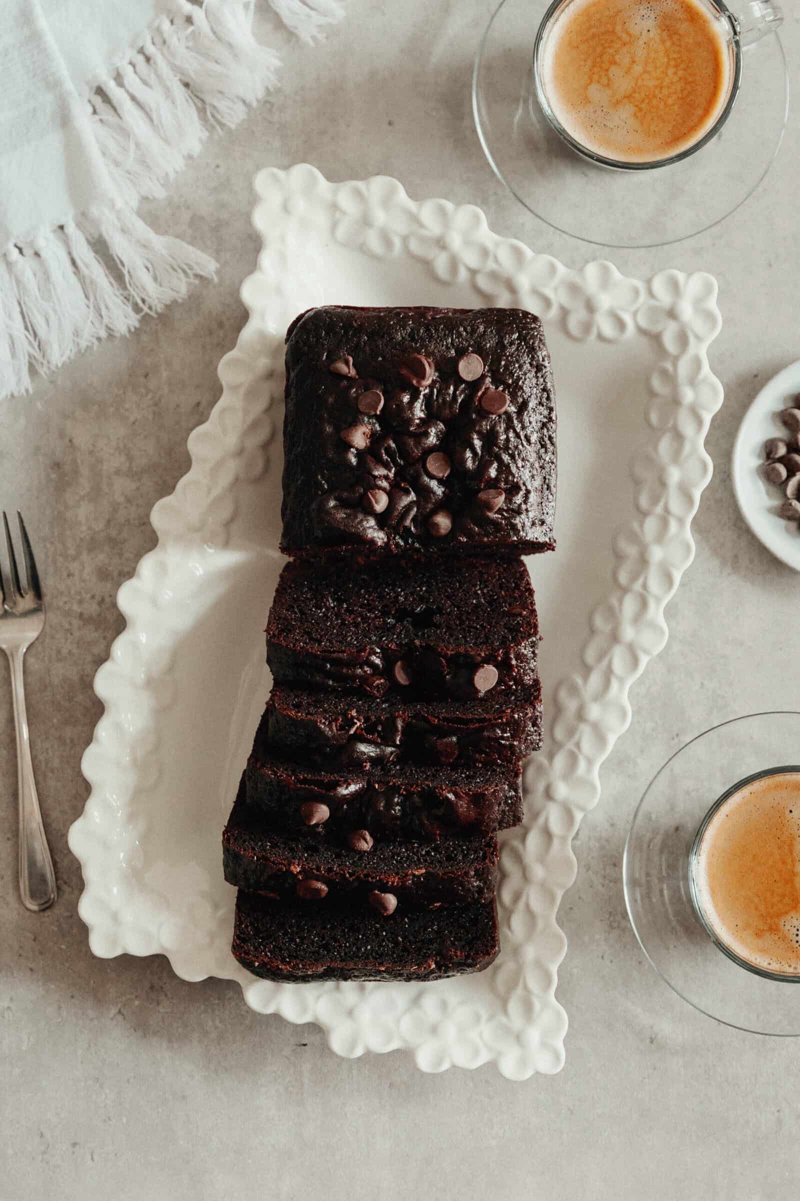 A sliced Chocolate Loaf Cake topped with chocolate chips is arranged on a decorative white platter, accompanied by two cups of espresso and a fork on a light-colored surface.