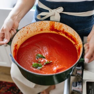 A person in a white top and blue apron holds a green pot filled with smooth tomato sauce, garnished with fresh basil leaves, standing in a kitchen by a stove.