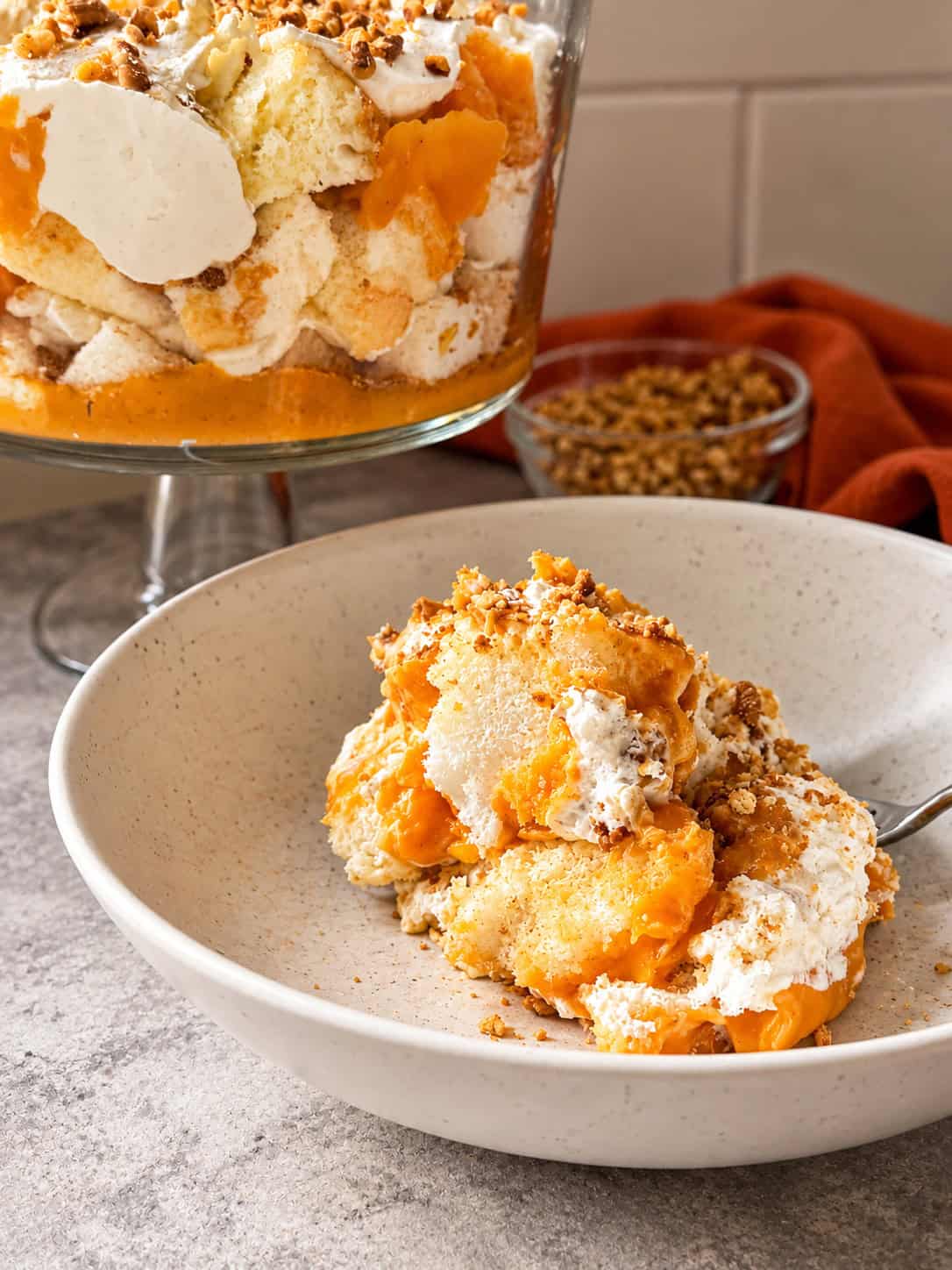 A serving of pumpkin trifle with layers of cake, whipped cream, and orange pumpkin filling in a ceramic bowl, with a large glass trifle dish in the background.