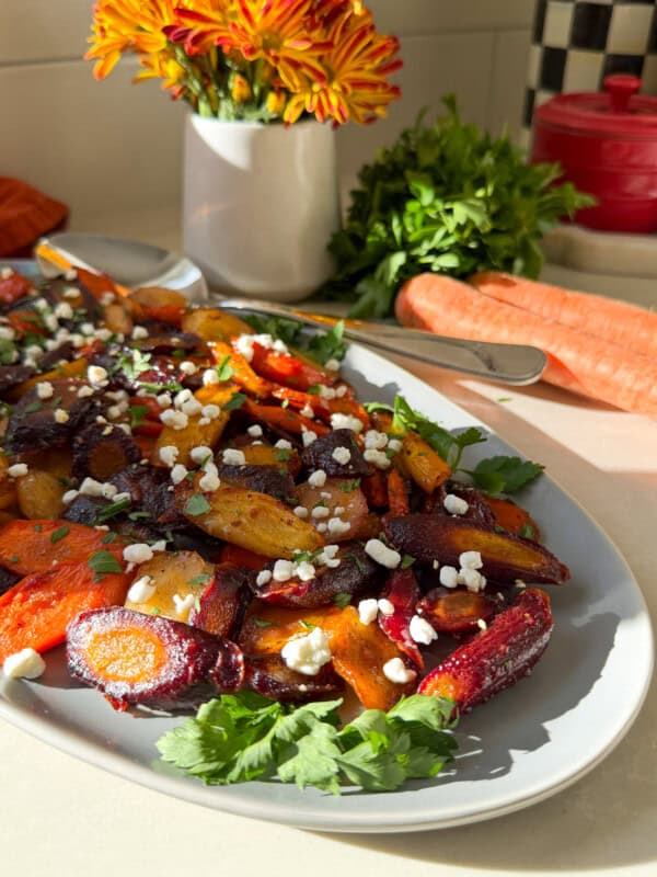 A plate of roasted carrots and beets, inspired by a Honey Carrots recipe, is topped with crumbled cheese and parsley on a sunlit countertop. In the background are fresh carrots, parsley, a red pot, orange flowers, and a checkered container.
