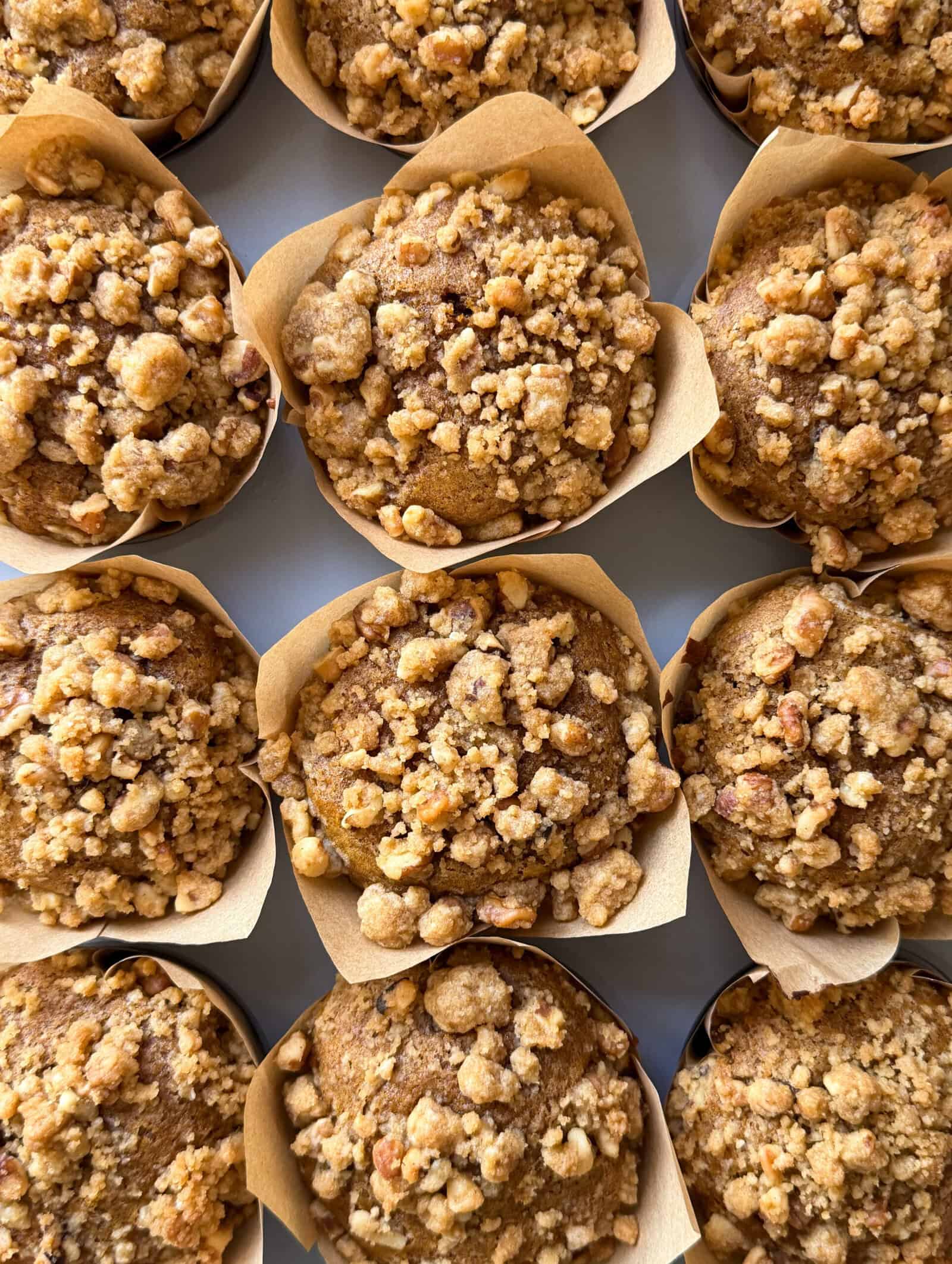 A close-up of several freshly baked Pumpkin Streusel Muffins topped with chunky brown sugar streusel, each in a tan paper liner, arranged closely together in neat rows.