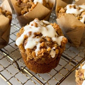 A close-up of Pumpkin Streusel Muffins topped with crumbly streusel and white icing, sitting on a wire cooling rack with more muffins in brown parchment wrappers in the background.