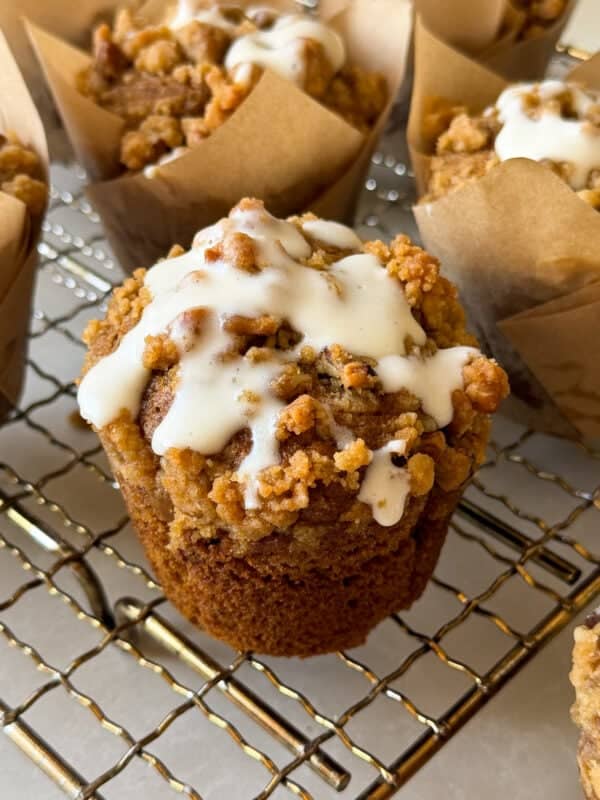 A close-up of Pumpkin Streusel Muffins topped with crumbly streusel and white icing, sitting on a wire cooling rack with more muffins in brown parchment wrappers in the background.
