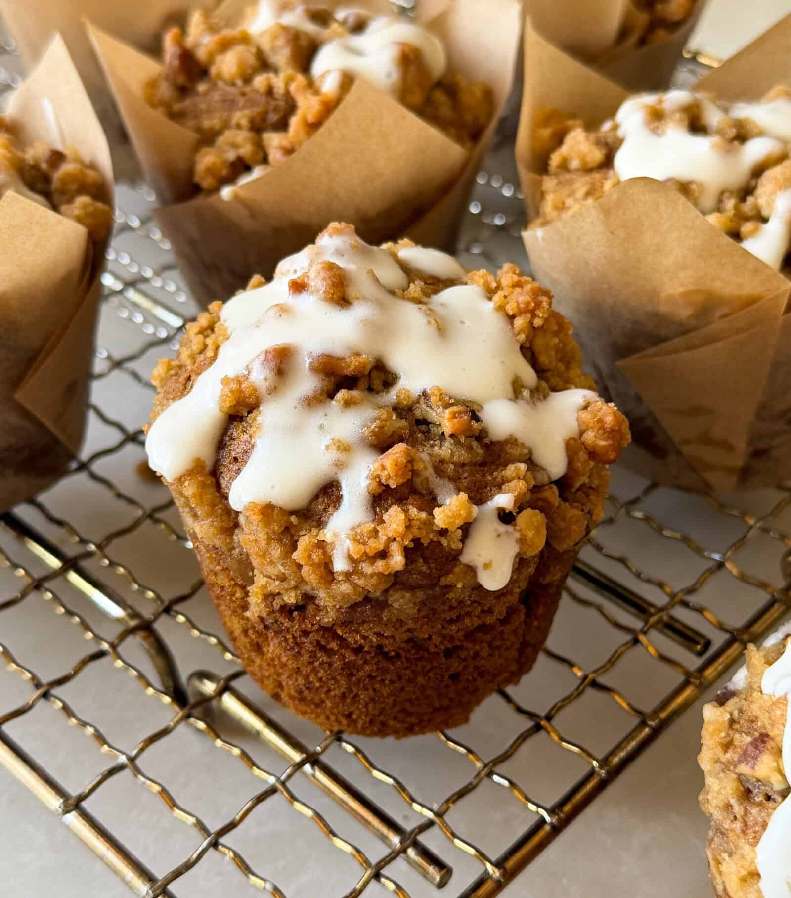 A freshly baked Pumpkin Streusel Muffin with crumb topping and white icing drizzle sits in a brown paper liner on a cooling rack, surrounded by other similar pumpkin muffins.
