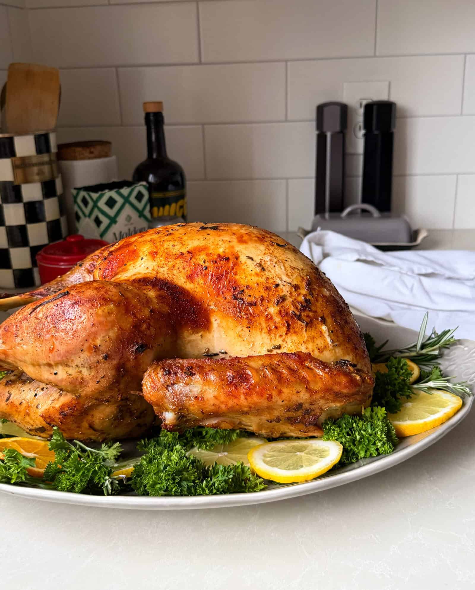 A golden-brown roasted turkey, prepared with a classic Roasted Turkey Recipe, is garnished with fresh parsley and lemon slices on a white platter atop the kitchen counter, surrounded by utensils and folded napkins in the background.