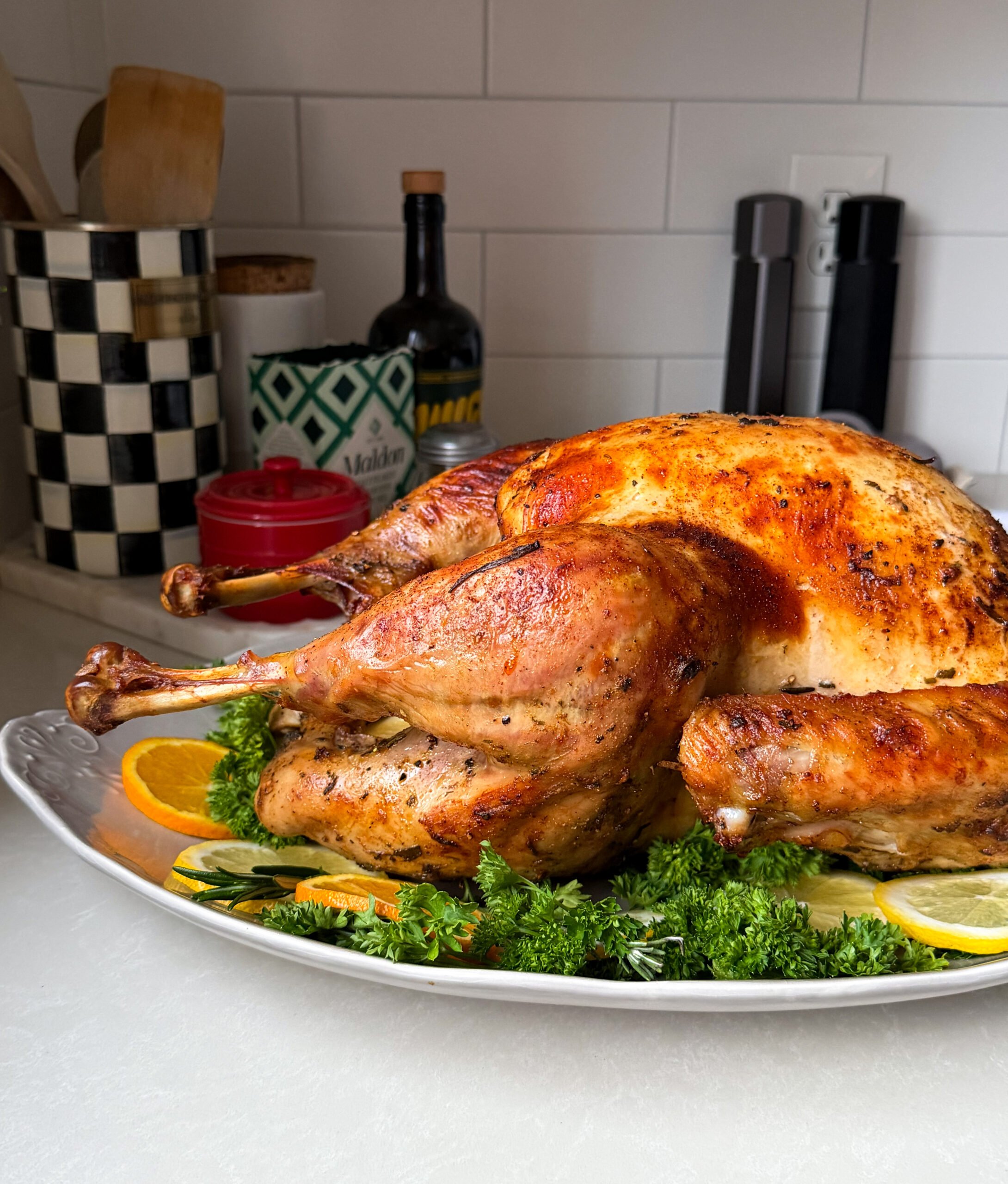 A roasted turkey, prepared using a classic Roasted Turkey Recipe, is garnished with fresh parsley and orange slices on a white platter atop the kitchen counter, with various kitchen items and a white tiled backsplash in the background.