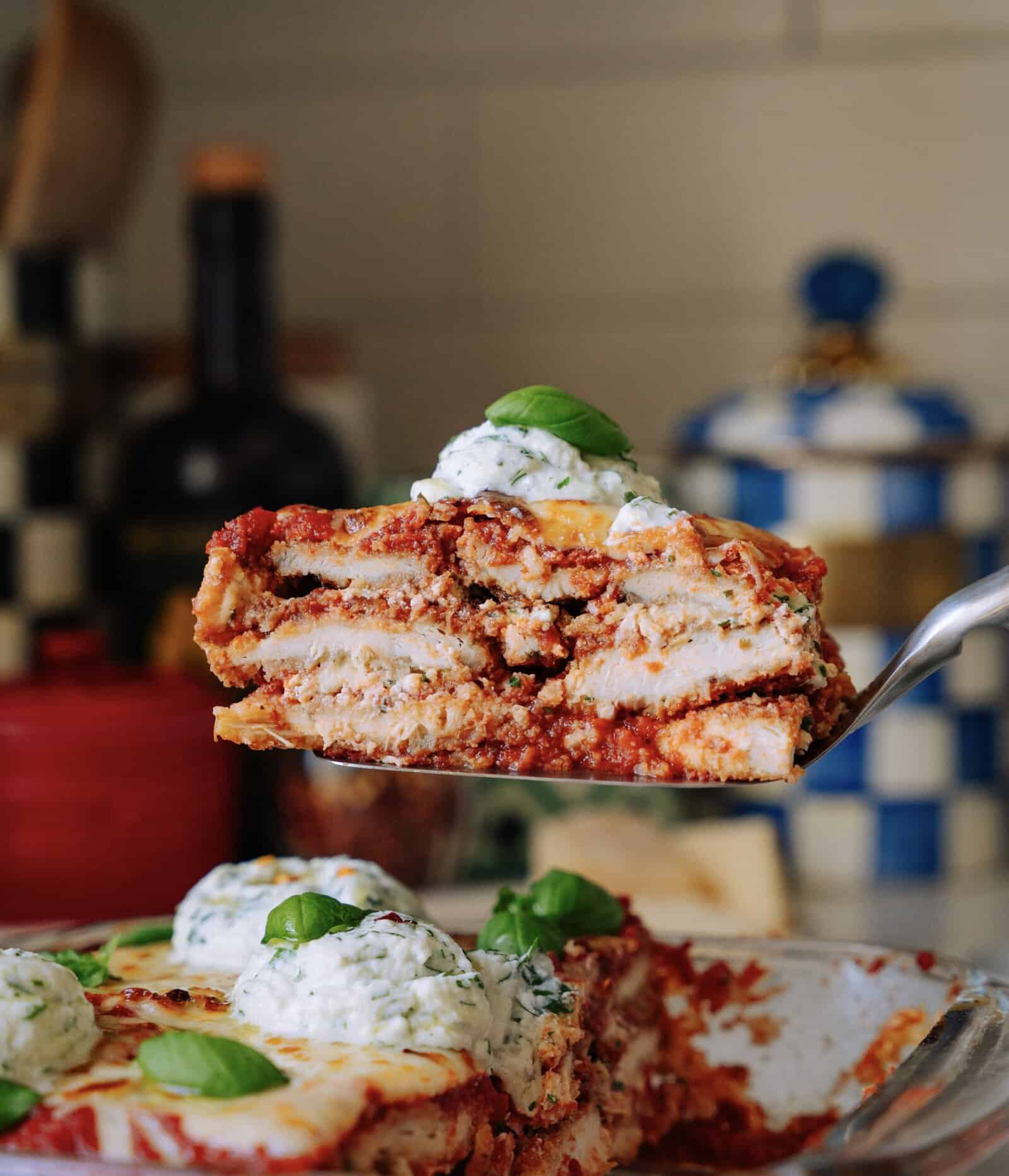 A close-up of a slice of homemade lasagna being lifted from a baking dish, topped with a scoop of ricotta cheese and a fresh basil leaf, with layers of sauce, pasta, and cheese visible.
