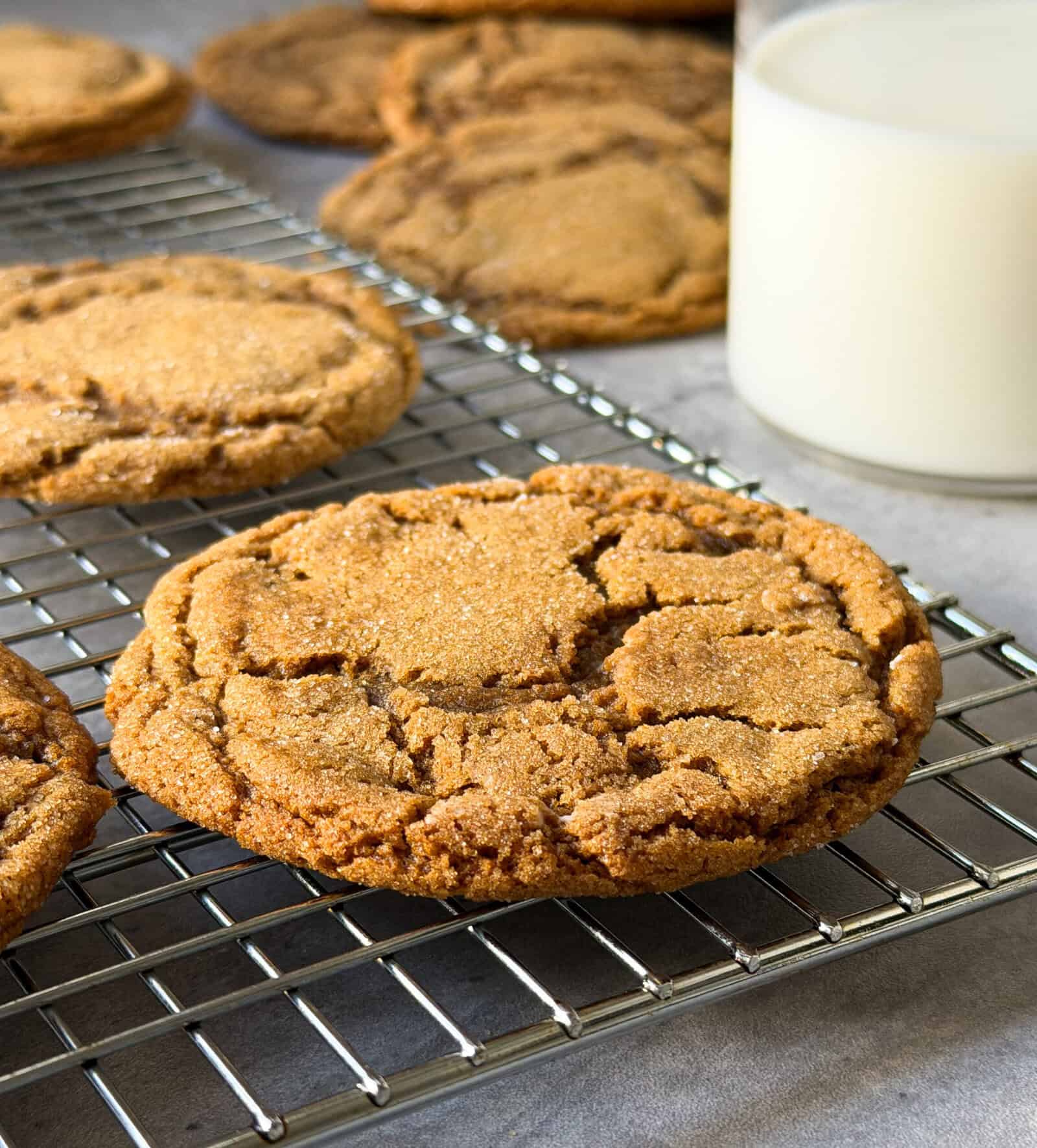 Close-up of a golden-brown Ginger Molasses Cookie on a cooling rack, with more Soft Gingerbread Cookies and a glass of milk in the background. The cookie’s surface is slightly cracked and sprinkled with sugar.