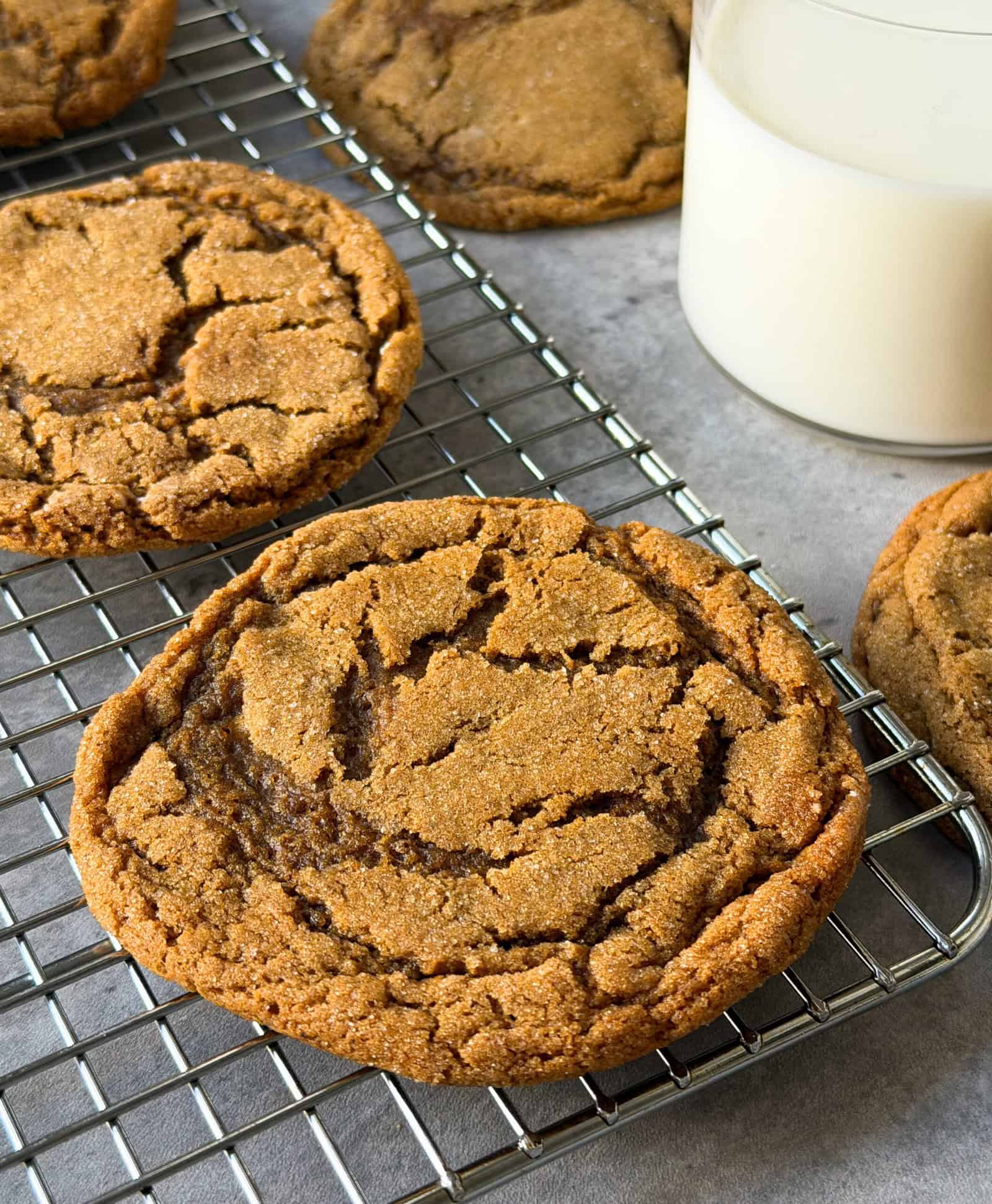 A close-up of large, golden brown Ginger Molasses Cookies with a cracked surface, cooling on a metal rack next to a glass of milk on a gray surface.