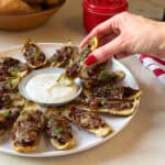 A hand with red nails picks up a loaded potato skin from a plate of cheesy, bacon-topped French Onion Potato Skins, arranged around a bowl of white dipping sauce—an irresistible twist on the classic Potato Skins Recipe.