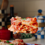 A fork holds a serving of baked ziti covered in tomato sauce and cheese above a casserole dish filled with more pasta, garnished with basil leaves, in a cozy kitchen setting.