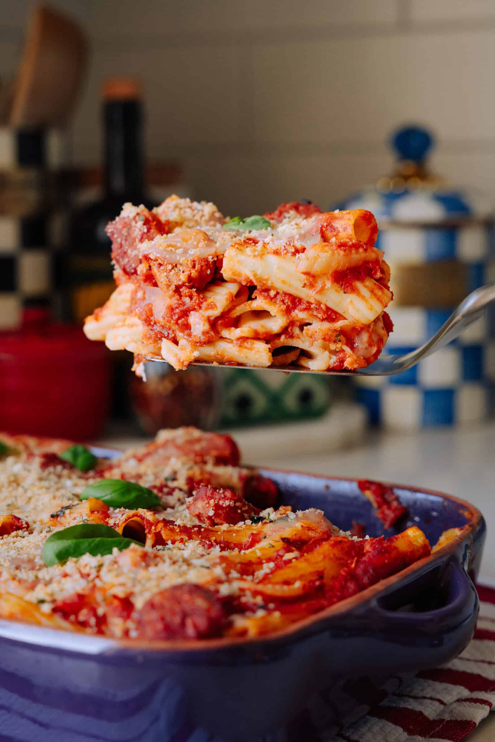 A close-up of baked ziti topped with tomato sauce, cheese, and basil being lifted from a purple baking dish with a spatula. The background features kitchen decor and containers.