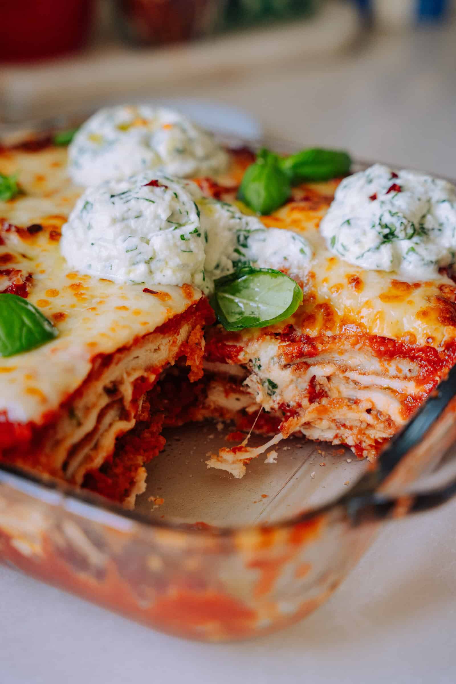 A glass baking dish of lasagna with a slice removed, showing layers of pasta, sauce, and cheese. The top is golden and bubbling, garnished with dollops of ricotta and fresh basil leaves.