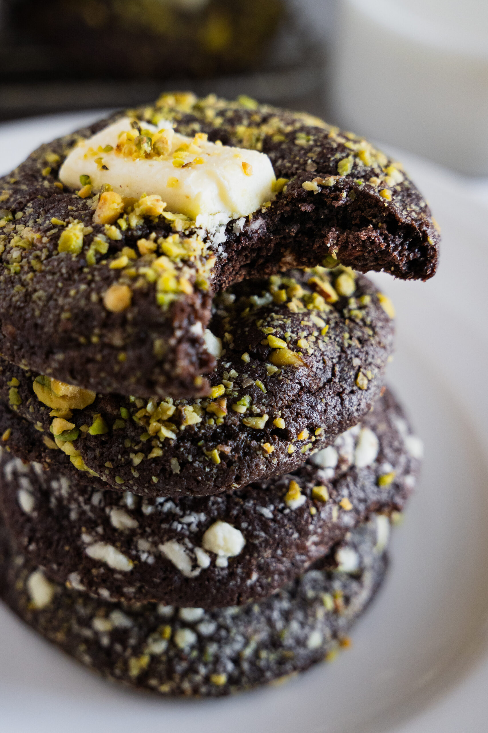 A stack of four chocolate cookies topped with chopped pistachios and white chocolate chunks sits on a white plate. The top cookie has a bite taken out and a piece of white chocolate on it.