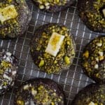 Chocolate cookies topped with crushed pistachios and small pieces of white chocolate, arranged on a metal cooling rack.