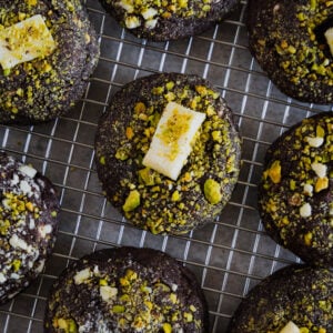 Chocolate cookies topped with crushed pistachios and small pieces of white chocolate, arranged on a metal cooling rack.