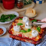 A glass baking dish of baked lasagna topped with scoops of herbed ricotta, surrounded by plates of fresh basil, grated Parmesan, red pepper flakes, and kitchen items on a countertop.