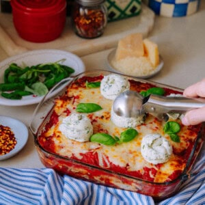 A glass baking dish of baked lasagna topped with scoops of herbed ricotta, surrounded by plates of fresh basil, grated Parmesan, red pepper flakes, and kitchen items on a countertop.