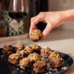 A hand holds a meatball above a dark plate filled with more meatballs, garnished with herbs and grated cheese. Inspired by the comfort of an Easy Stuffed Mushrooms recipe, red wine and kitchen items sit in the background on the countertop.