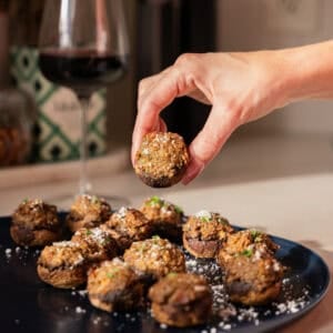 A hand holds a meatball above a dark plate filled with more meatballs, garnished with herbs and grated cheese. Inspired by the comfort of an Easy Stuffed Mushrooms recipe, red wine and kitchen items sit in the background on the countertop.