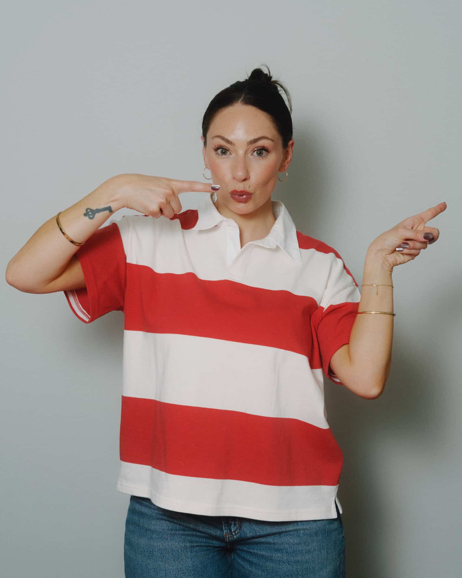 A woman in a red and white striped shirt and jeans points at her cheek with one hand and gestures sideways with the other, standing against a plain light gray background.