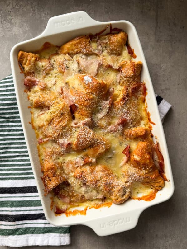 A baked casserole in a white rectangular dish, inspired by a Ham and cheese croissant bake, is topped with melted cheese and pieces of ham, resting on a striped kitchen towel on a gray surface.