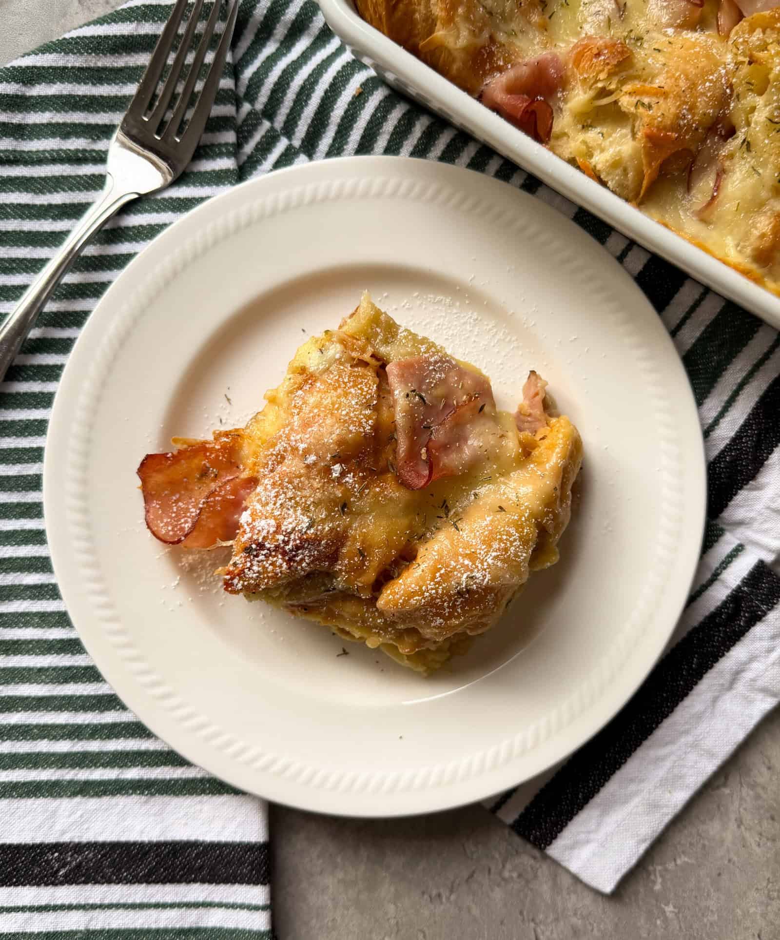 A slice of savory Ham and cheese croissant bake rests on a white plate beside a baking dish. A fork sits on a green and white striped towel—perfect for your Christmas morning breakfast.