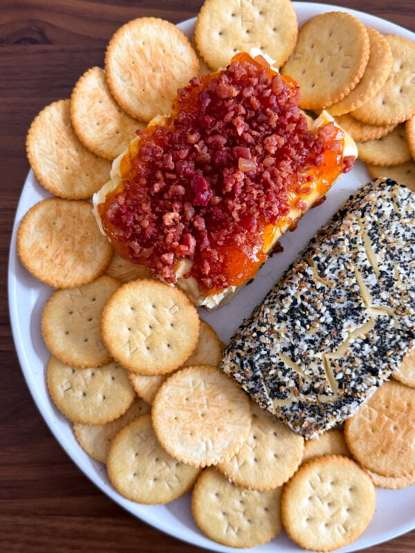 A plate of round crackers surrounds two cheese spreads: one is a baked cream cheese topped with bacon and red sauce, while the other is coated in black and white seeds with shredded cheese on top.