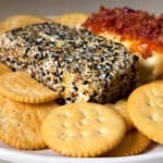 A close-up of a plate with round butter crackers, a rectangular block of baked cream cheese coated in everything bagel seasoning, and another cheese block topped with crumbled bacon and melted cheese.
