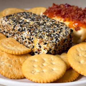 A close-up of a plate with round butter crackers, a rectangular block of baked cream cheese coated in everything bagel seasoning, and another cheese block topped with crumbled bacon and melted cheese.