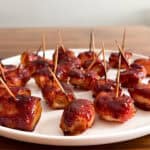 A white plate with bite-sized bacon wrapped water chestnuts, glazed with a shiny sauce and secured with toothpicks, set on a wooden table—a classic bacon wrapped water chestnuts recipe.