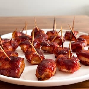 A white plate with bite-sized bacon wrapped water chestnuts, glazed with a shiny sauce and secured with toothpicks, set on a wooden table—a classic bacon wrapped water chestnuts recipe.