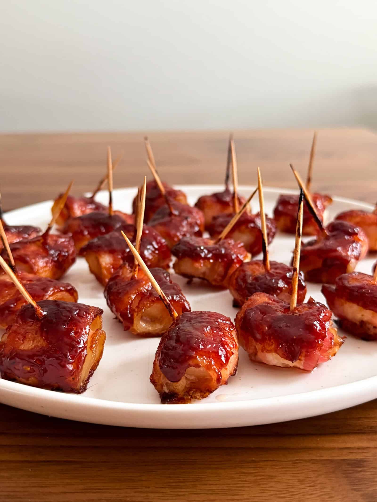 A white plate with bite-sized bacon wrapped water chestnuts, glazed with a shiny sauce and secured with toothpicks, set on a wooden table—a classic bacon wrapped water chestnuts recipe.