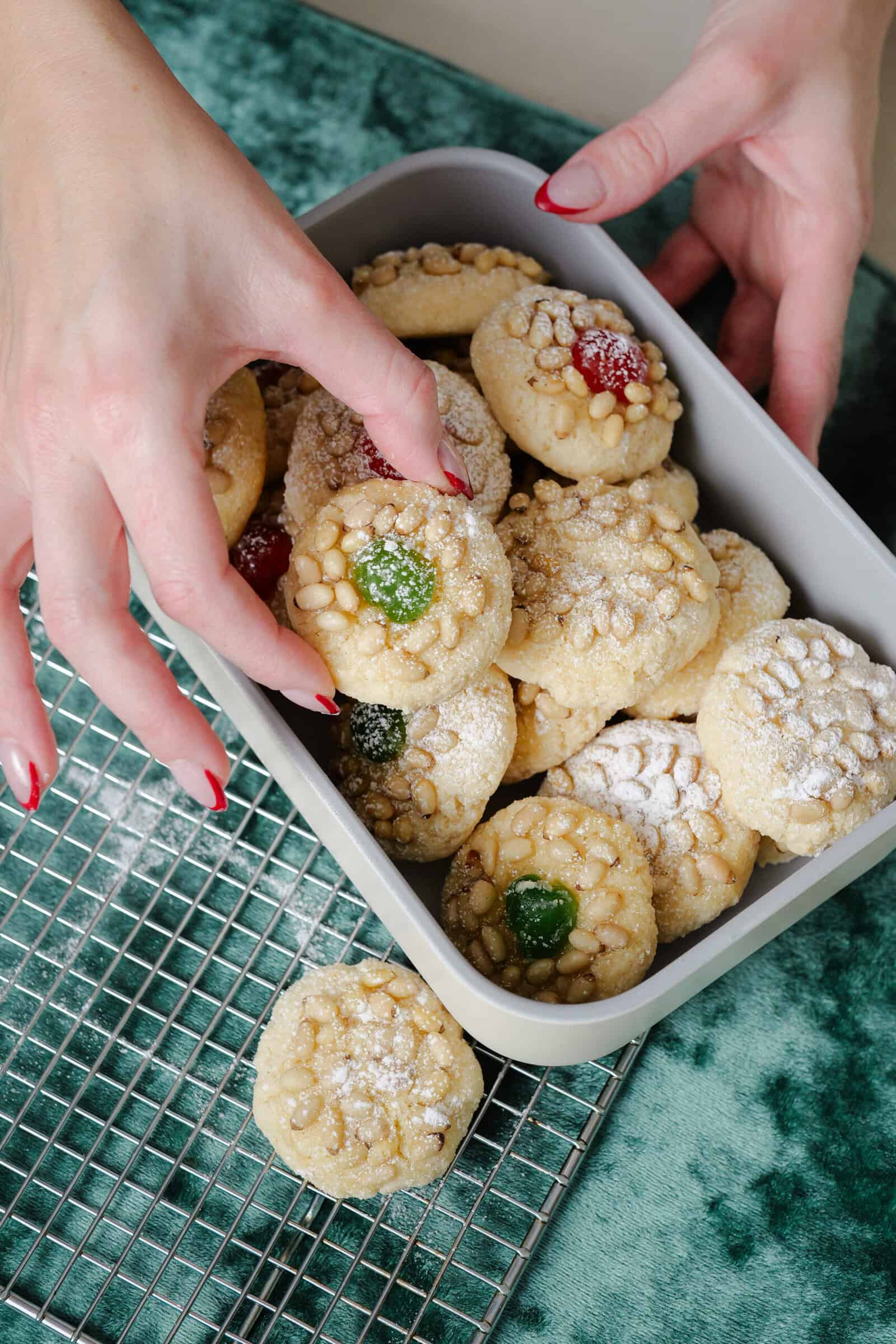 A person with red-painted nails places a round cookie topped with a red candied cherry into a container filled with similar cookies, some topped with green cherries and dusted with powdered sugar, on a cooling rack and green fabric.