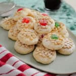A plate of pine nut cookies dusted with powdered sugar, some topped with red or green candied cherries, sits on a table with a red-and-white striped cloth and a glass cup of espresso in the background.