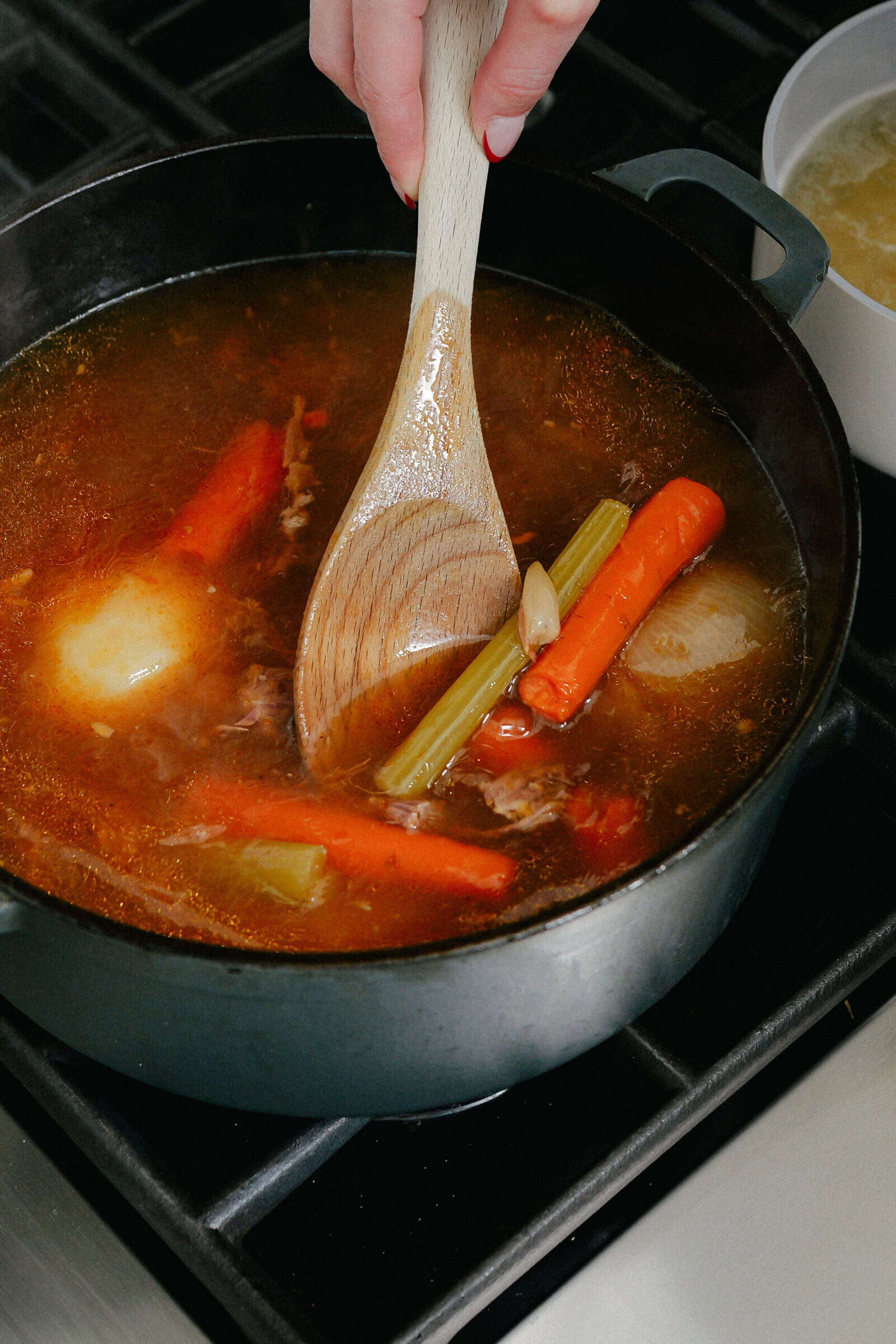 A hand stirs a pot of simmering broth with a wooden spoon; visible ingredients include carrots, celery, onions, and garlic. The pot sits on a stovetop next to a bowl of noodles.