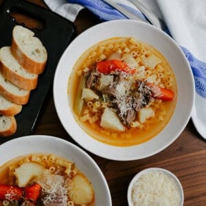 Two bowls of soup with pasta, shredded meat, carrots, celery, and potatoes, topped with grated cheese. Nearby are a small bowl of extra cheese, sliced baguette on a black tray, and two spoons on a blue-striped towel.
