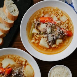 Two bowls of soup with pasta, shredded beef, carrots, celery, and potatoes. Grated cheese is sprinkled on top. Slices of baguette are on a cutting board, with extra grated cheese in a small bowl nearby.