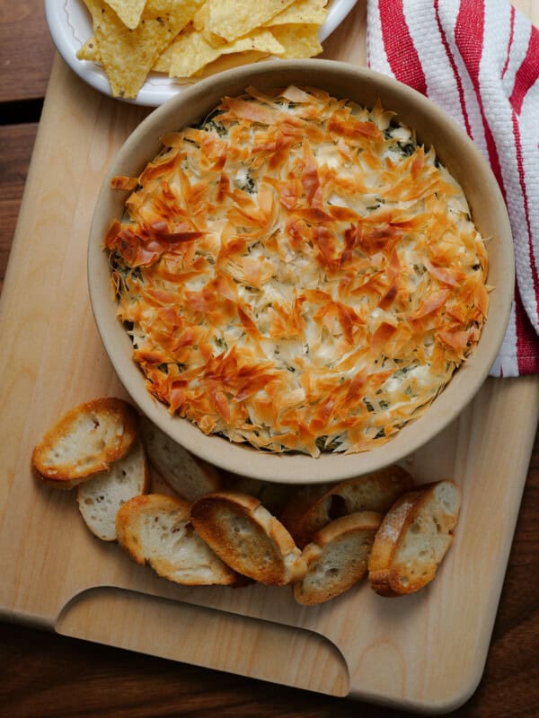 A round baking dish filled with a golden, flaky-topped dip sits on a wooden board, surrounded by toasted baguette slices. A bowl of tortilla chips and a red-striped kitchen towel are also visible.