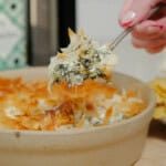 A hand holding a spoon lifts a serving of spinach and cheese dip with crispy phyllo pastry topping from a round beige baking dish.