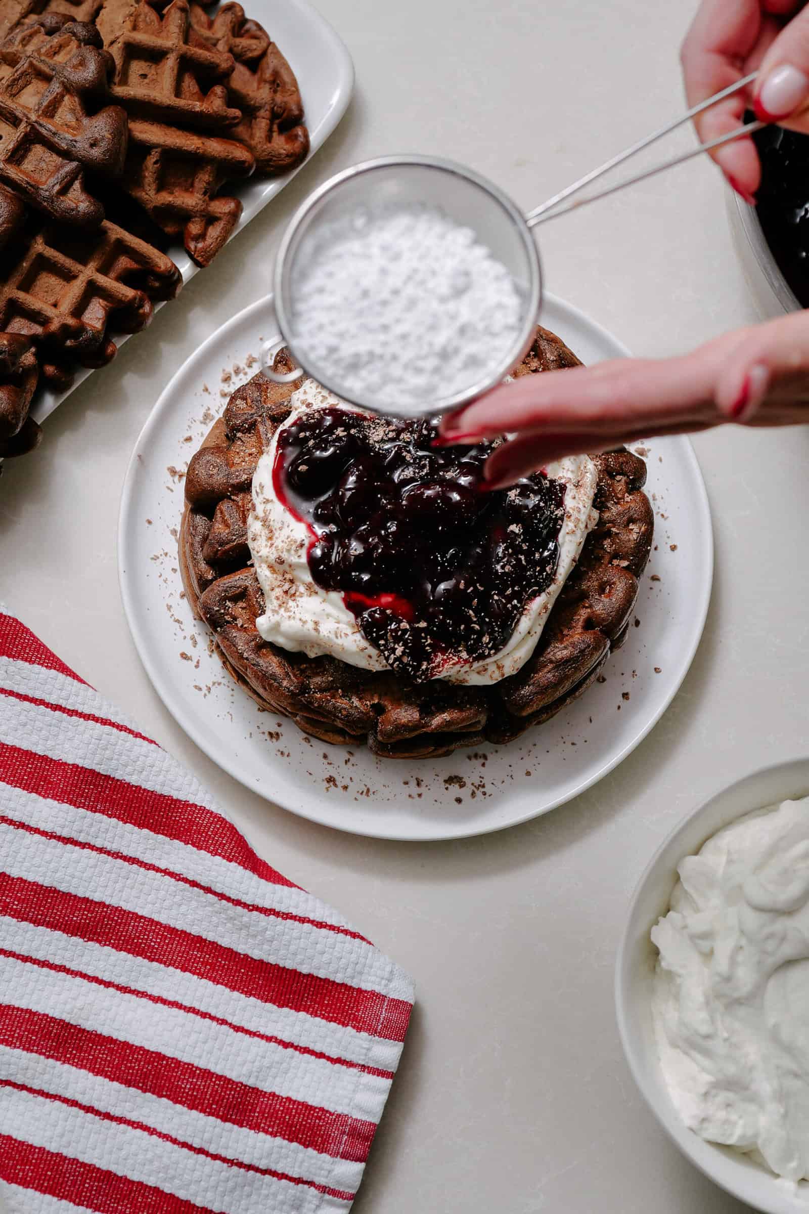 A hand sprinkles powdered sugar over chocolate waffles topped with whipped cream and berry sauce on a white plate. Nearby are more waffles, a bowl of whipped cream, and a red-striped cloth.