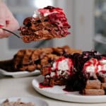 A hand lifts a slice of chocolate waffles topped with whipped cream and berry sauce from a plate stacked with more waffles, with extra waffles and a bowl of chocolate shavings in the background.