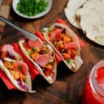 Three soft tacos filled with seasoned chicken, pickled onions, shredded cabbage, and sliced green onions are displayed on a wooden board. A small plate of chopped green onions and extra tortillas are in the background.