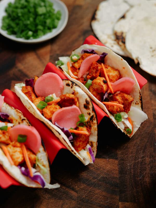 Three soft tacos filled with grilled chicken, pickled onions, shredded carrots, cabbage, and green onions are displayed in a red taco holder on a wooden surface, with a small plate of chopped herbs and tortillas in the background.