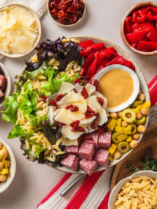 A salad bowl with mixed greens, orzo pasta, salami cubes, shaved parmesan, olives, chickpeas, red peppers, sun-dried tomatoes, and dressing, surrounded by small bowls of salad toppings on a light table.