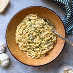 A bowl of creamy garlic pasta garnished with chopped herbs sits on a gray surface, with a fork in the dish. Nearby are a wedge of cheese, garlic cloves, and a striped kitchen towel—perfect inspiration for your next Garlic Pasta Recipe.